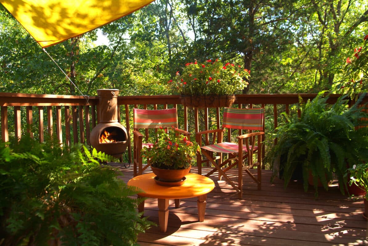 Horizontal image of an inviting outdoor sitting area surrounded by nature. A sun sail shades the porch and a chiminea burns pinion in the corner. Beautiful pants in containers adorn the outdoor room.